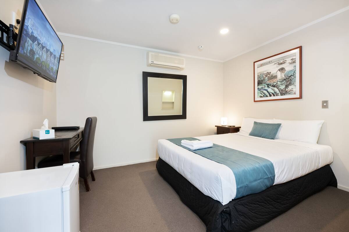 A hotel room with a bed, a desk, a chair, a television, an air conditioner, a framed picture, and a bedside table with a lamp.