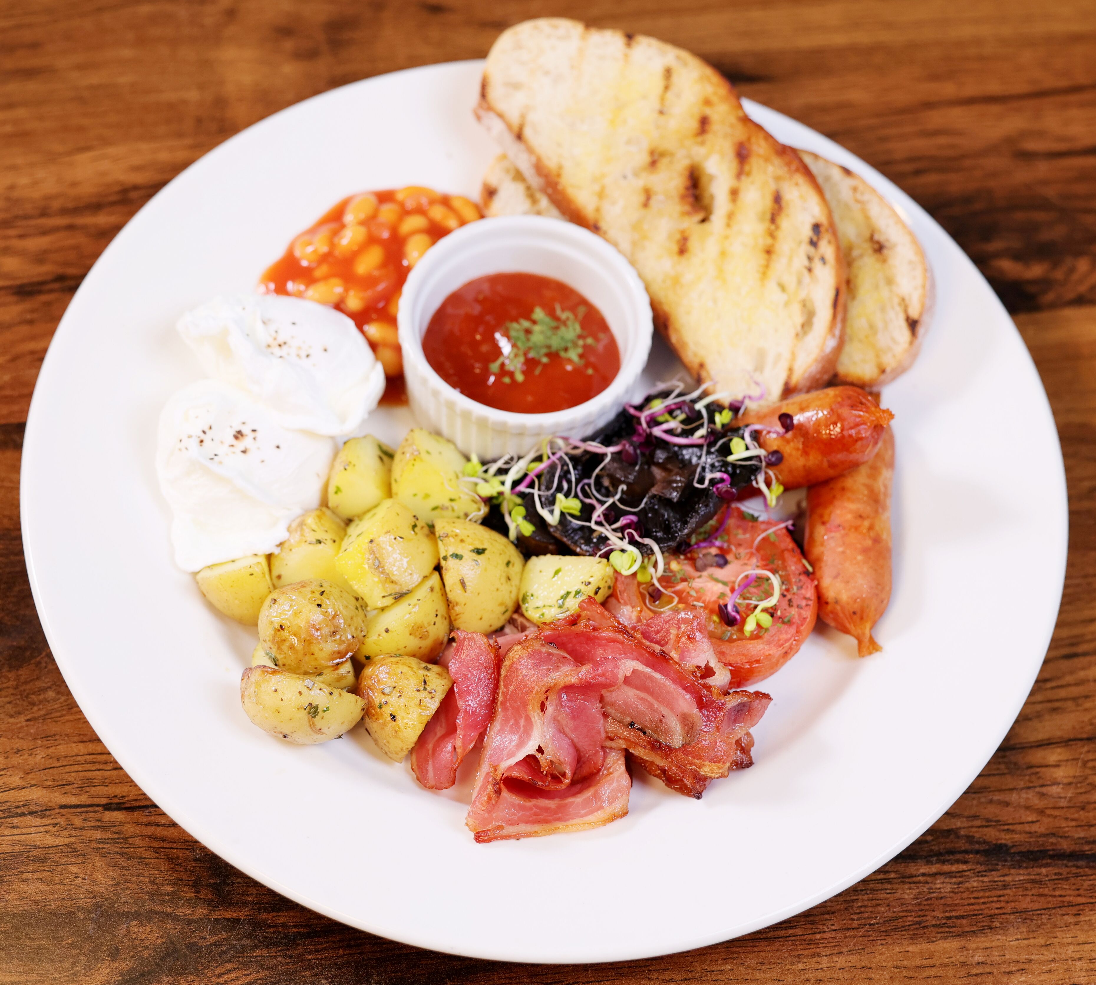 A plate with roasted potatoes, bacon, a poached egg, grilled tomatoes, and a side of beans.