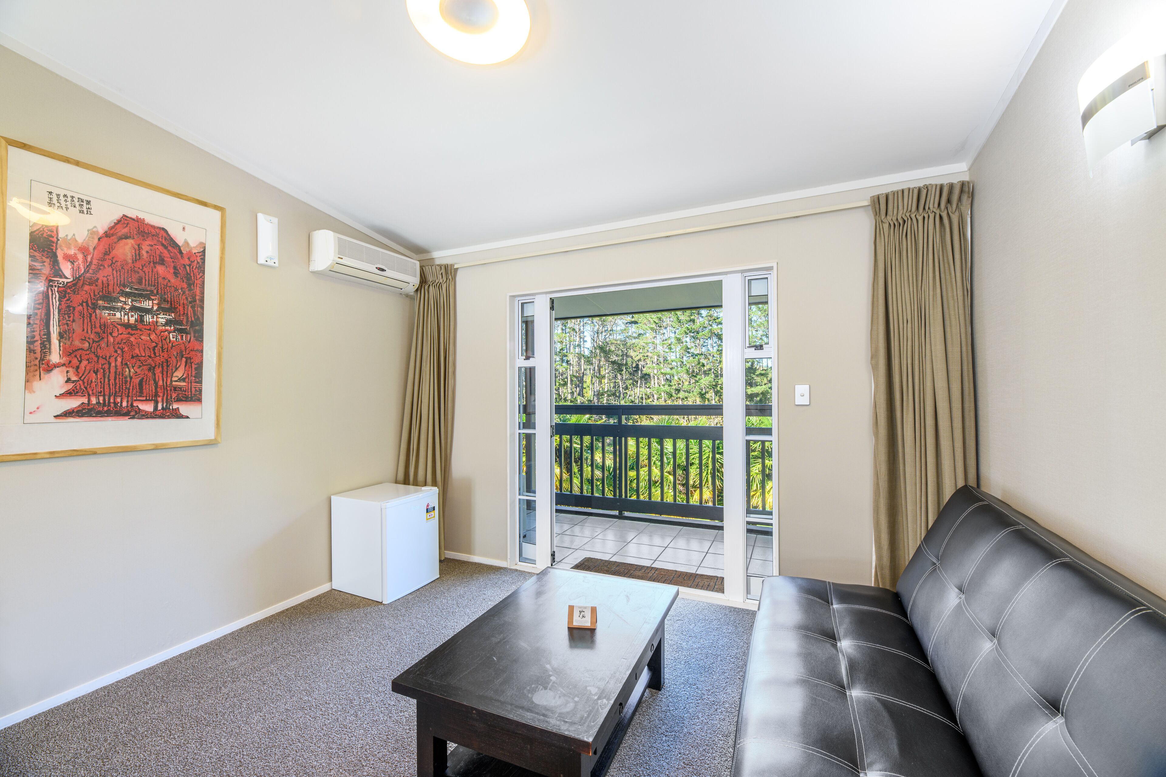 A living room with a sofa, a coffee table, a framed artwork, an air conditioning unit, and a sliding glass door leading to a balcony.