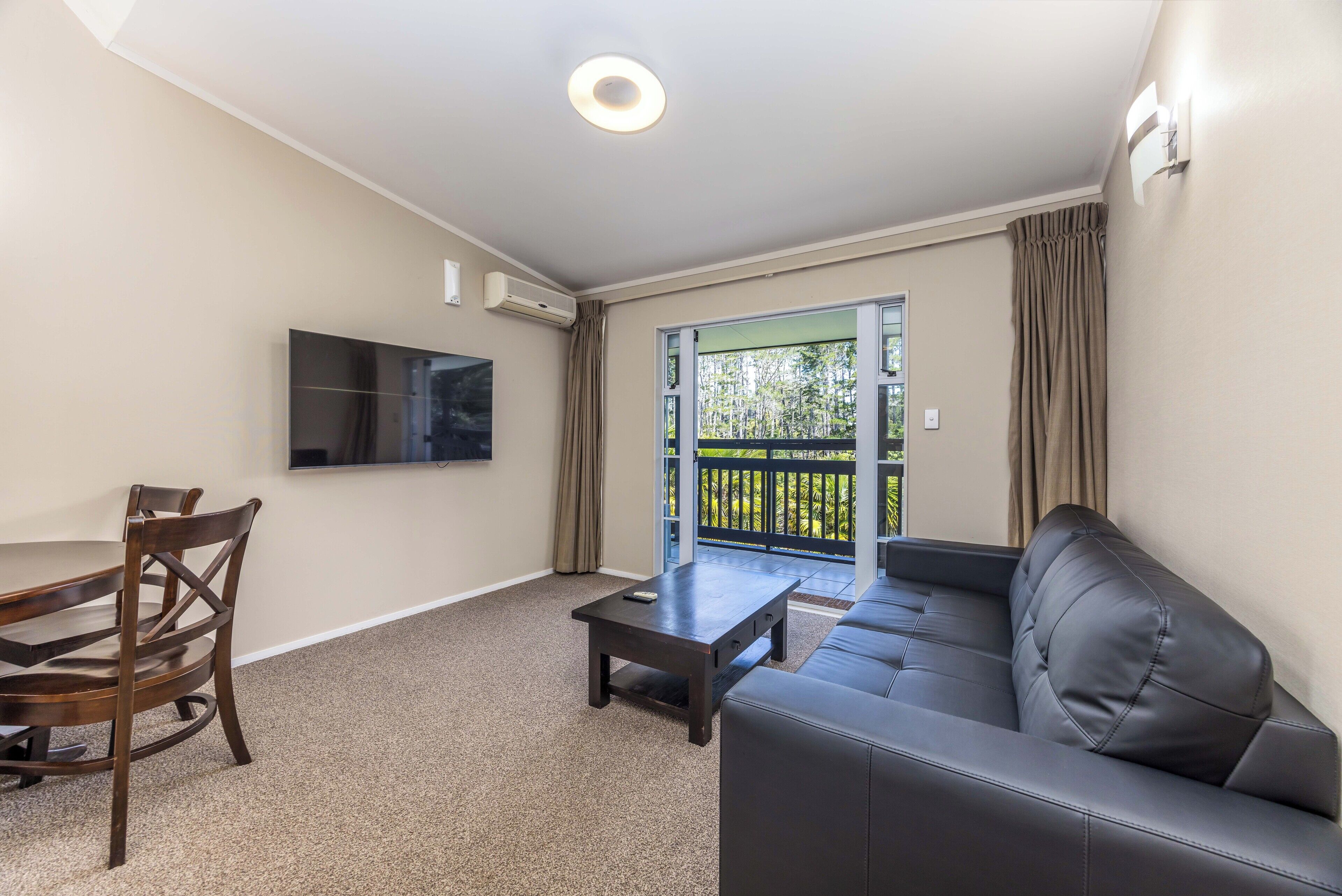 A living room with a sofa, a coffee table, a TV mounted on the wall, and a sliding glass door leading to an outdoor balcony.