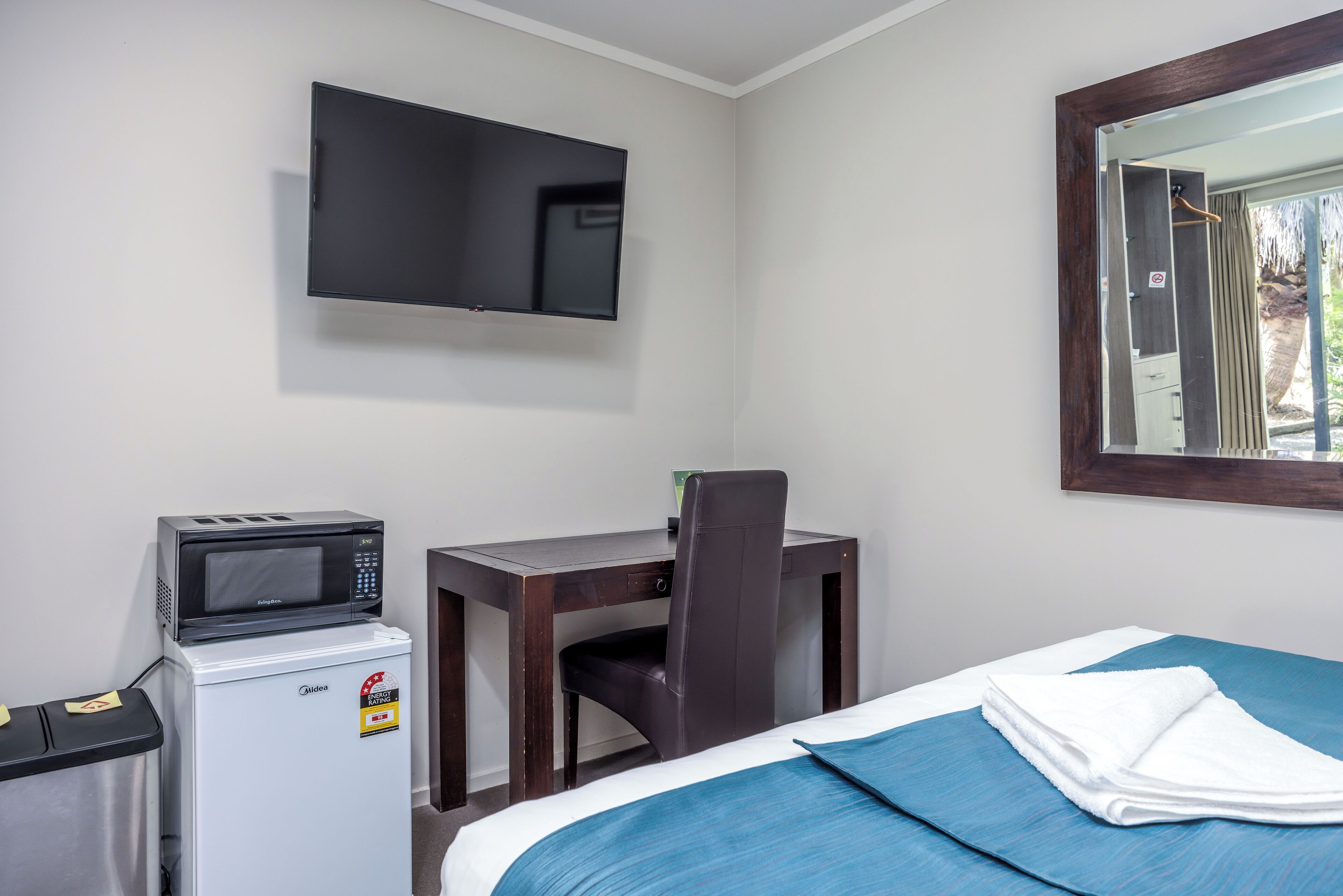 Ett hotellrum med en säng, ett skrivbord med en stol, en tv som är monterad på väggen, en mikrovågsugn och ett litet kylskåp.