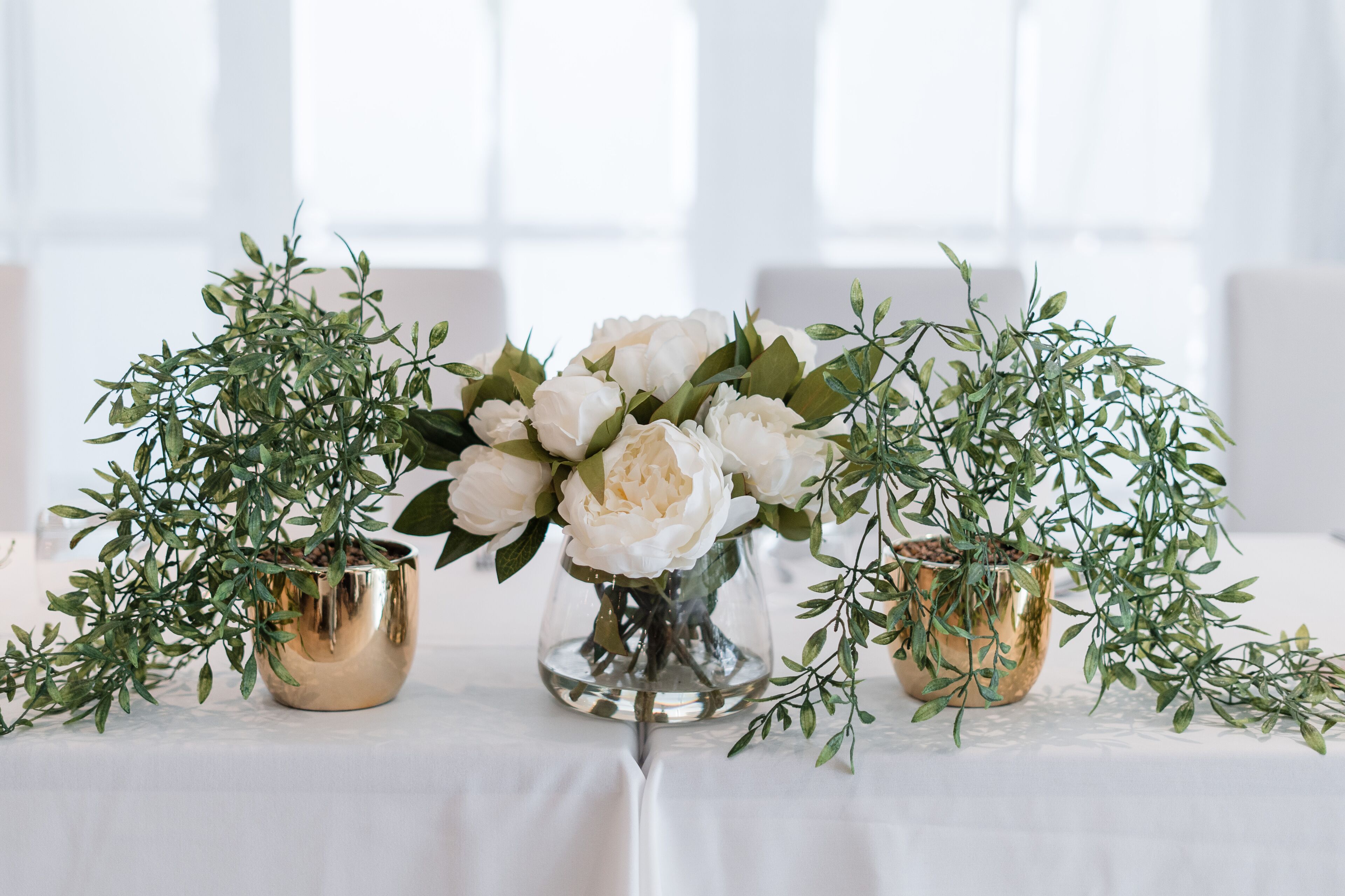 A table with three gold vases containing white flowers and green foliage.