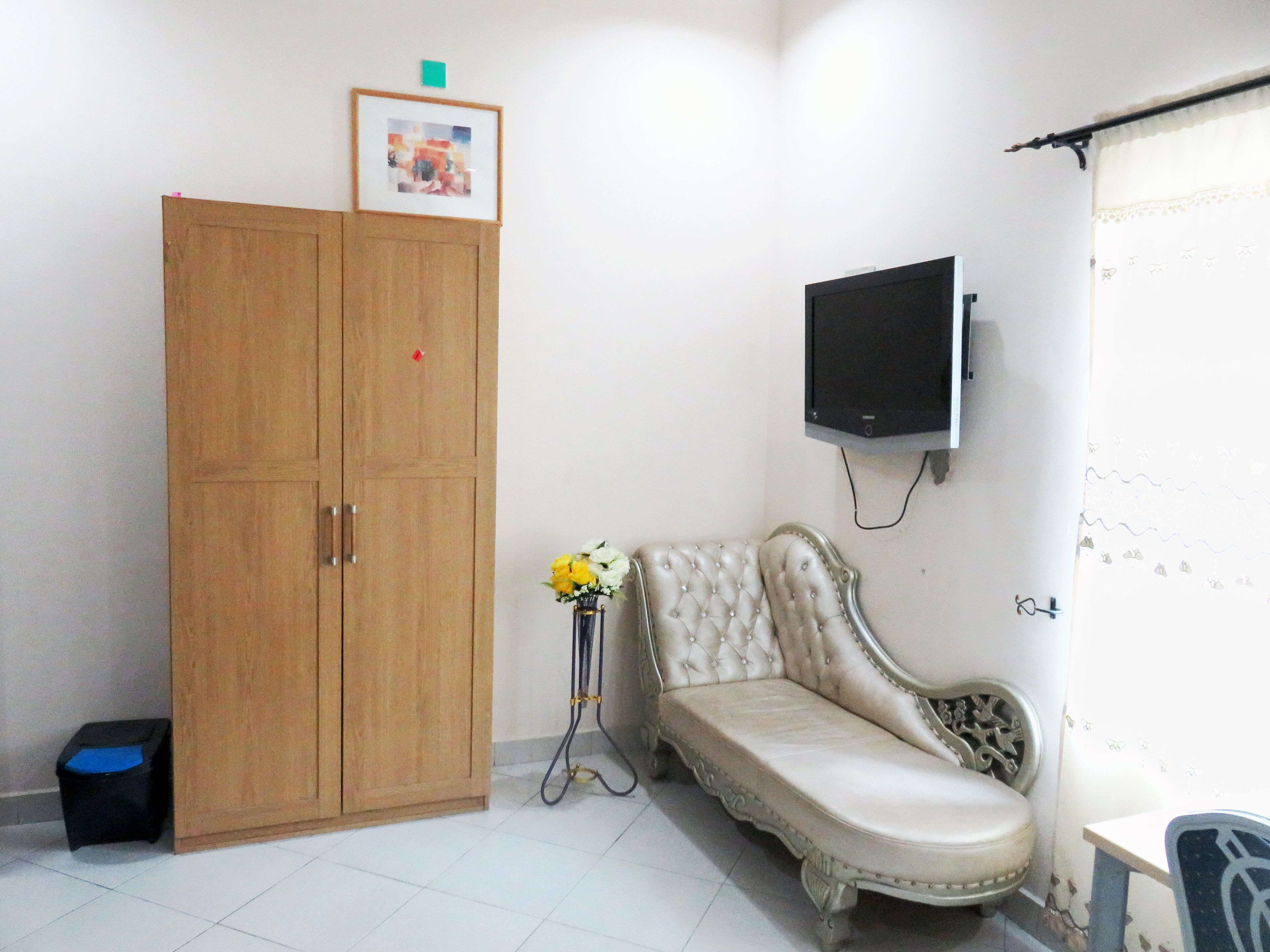 A room with a wooden wardrobe, a cushioned bench, a small table with a vase, and a mounted television.