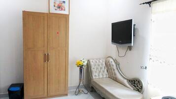A room with a wooden wardrobe, a cushioned bench, a small table with a vase, and a mounted television.