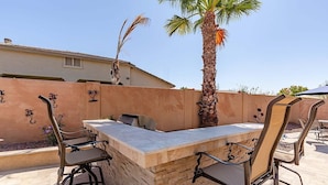 An outdoor bar area with a stone counter, metal chairs, and a palm tree. - Westgate Wonder ~ A Resort-Style Oasis w/ Heated Pool, Spa, Game Rooms, Putting Green & More! (Glendale)