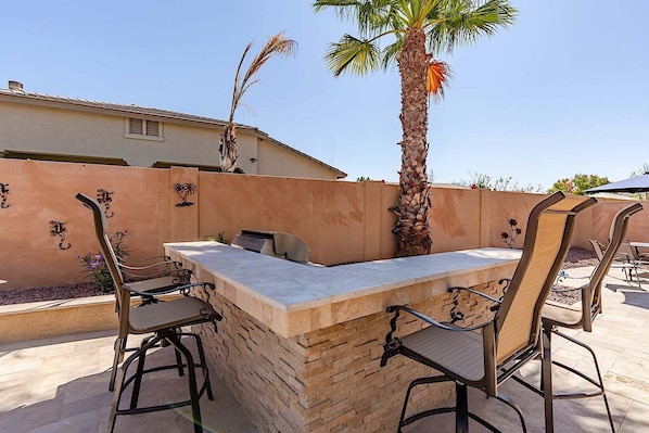 An outdoor bar area with a stone counter, metal chairs, and a palm tree. - Westgate Wonder ~ A Resort-Style Oasis w/ Heated Pool, Spa, Game Rooms, Putting Green & More! (Glendale)