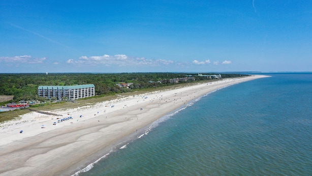 Eine Luftaufnahme eines Strandes mit einem großen Gebäude, sandigem Ufer und ruhigem Wasser.