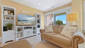A living room with a beige couch, a flat-screen TV, a bookshelf, and a view of the outdoors. - Oceanfront Sea Cloisters Family 3BR Villa • Private Stairs to Beach, Pool & Boardwalk (Hilton Head Island)