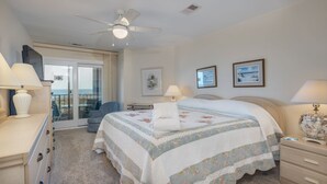 A hotel room with a bed, a desk, a chair, a ceiling fan, and a view of the sea through a sliding door. - Beachfront Sea Cloisters Villa w Ocean Views, Balcony & Complex Pool (Hilton Head Island)