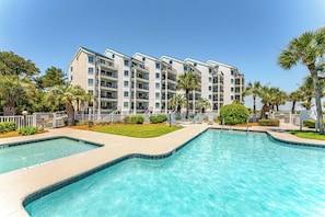 A multi-story hotel with a pool area, including two separate swimming pools and a landscaped garden with palm trees.