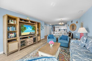 A living room with a television, wooden entertainment unit, blue sofas, and a kitchen in the background.