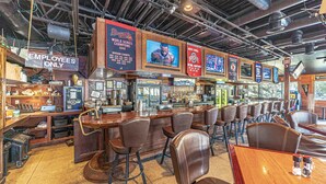 Eine Bar mit verschiedenen Sitzmöglichkeiten, darunter Barhocker und Tische. Mehrere Fernseher an den Wänden zeigen Sportübertragungen. Die Theke ist mit diversen Geräten und einer Kasse ausgestattet.