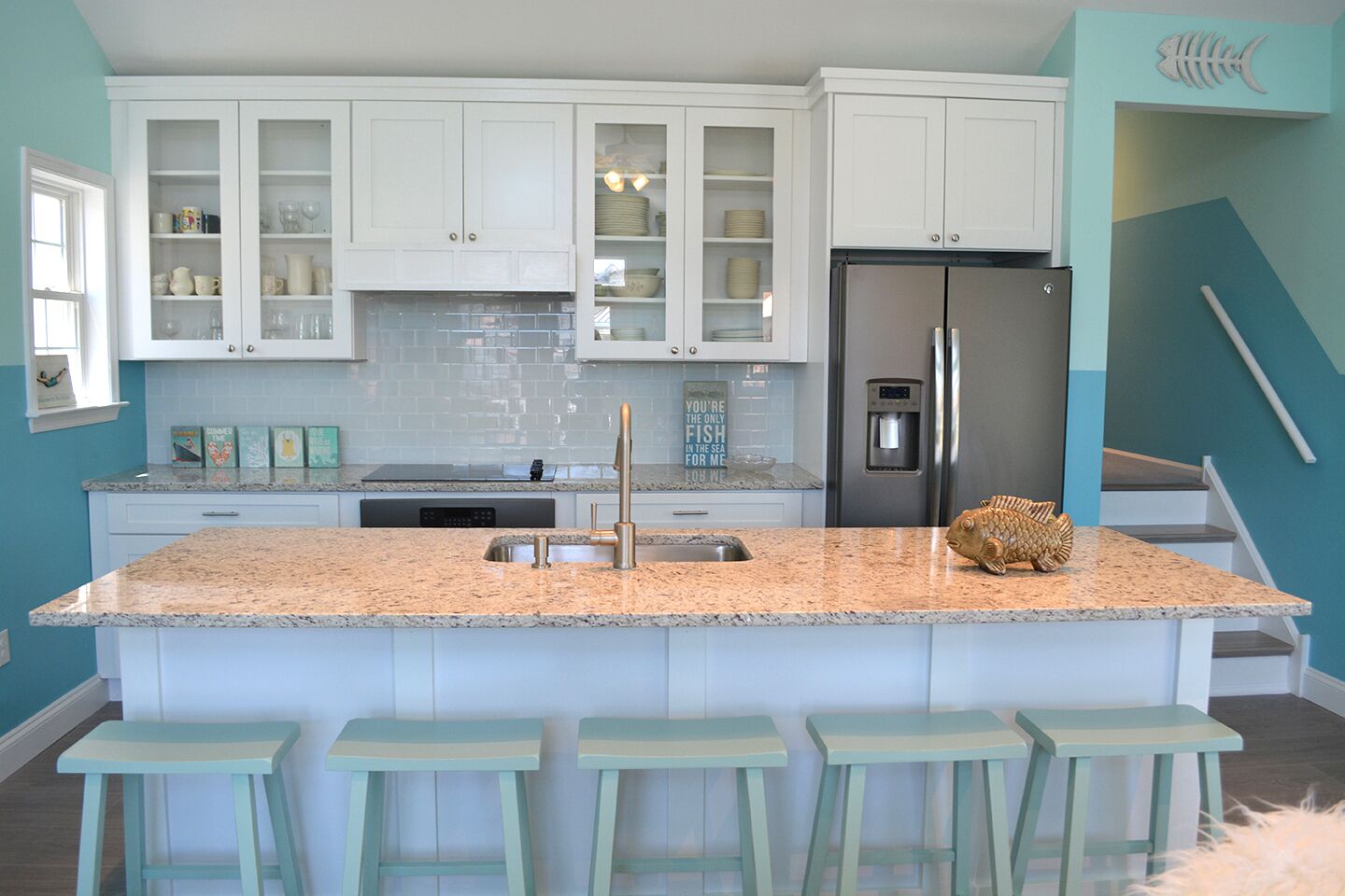 A kitchen with white cabinets, a stainless steel refrigerator, a granite countertop, and four light blue bar stools.