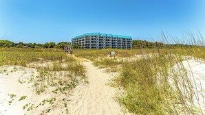 Et hotel ved stranden med en sandsti, der fører til indgangen, omgivet af højt græs og en klar blå himmel.