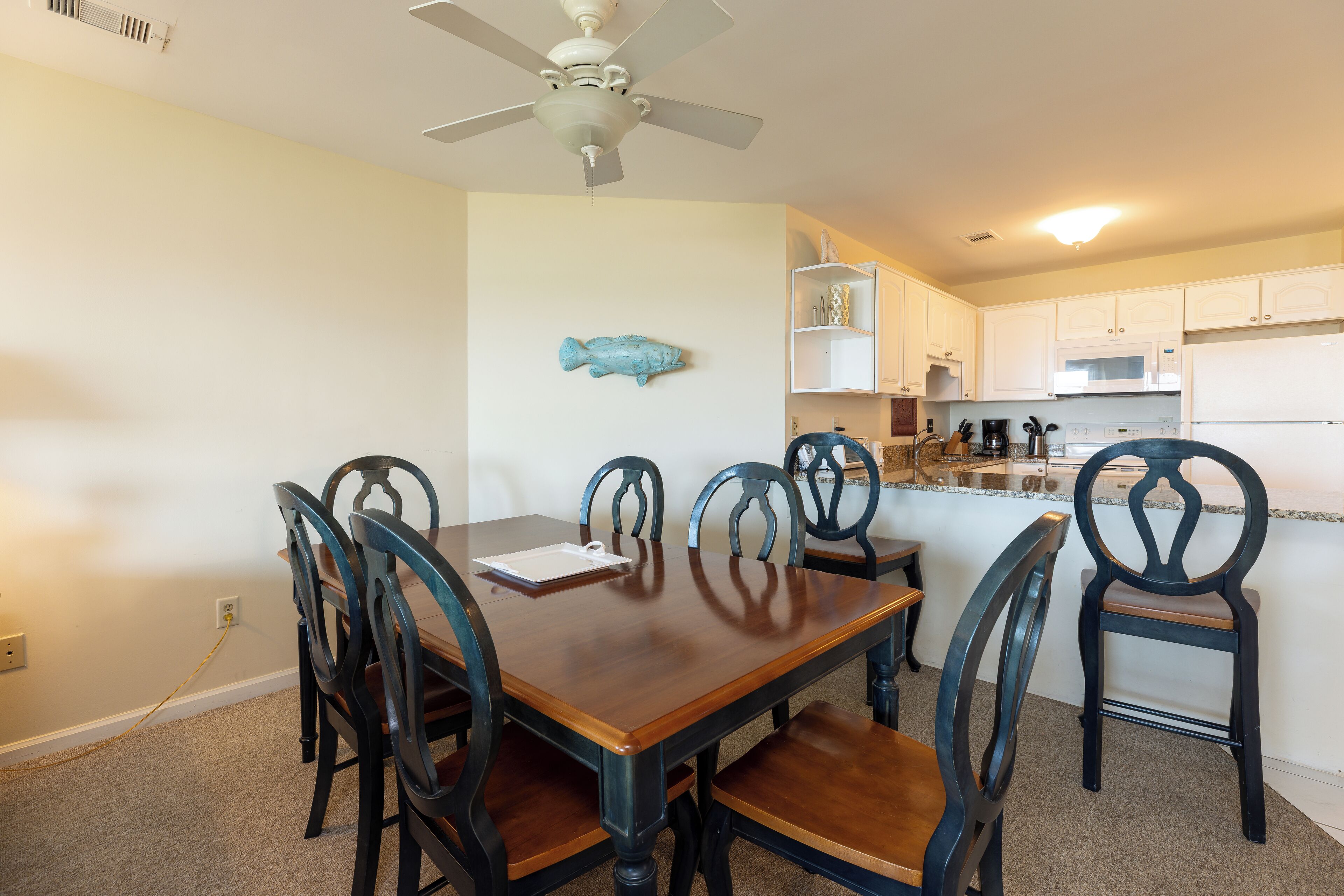 Een eethoek met een houten tafel en stoelen, een plafondventilator en witte keukenkastjes op de achtergrond.