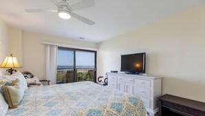 Ein Schlafzimmer mit Bett, Fernseher auf einer Kommode, Deckenventilator und Meerblick durch ein großes Fenster.
