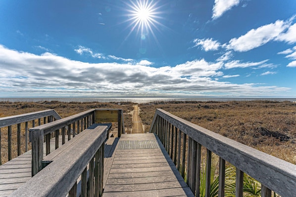 A wooden boardwalk leading to a beach with a clear sky and sun.