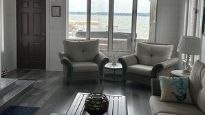 Una sala de estar con muebles de mimbre, una mesita redonda y vistas a una masa de agua a través de amplias ventanas.