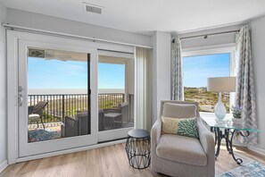 Una habitación con puerta corrediza de vidrio que da a un balcón, un sofá, una mesita de noche con lámpara y vistas al paisaje.