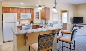 A compact kitchen with wooden cabinets, a granite countertop, a built-in microwave, a stove, and a sink. There are two bar stools with patterned upholstery. - Rare Find! 3 BR Dlx Sleeps 10 -Bonnet Creek Resort-5 Pools & More (Orlando)