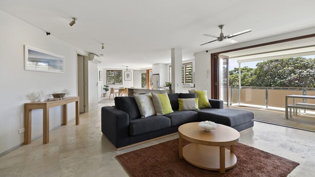 Uma sala de estar moderna com um sofá escuro, uma mesa de centro redonda de madeira e vista para o exterior através de grandes portas de correr.