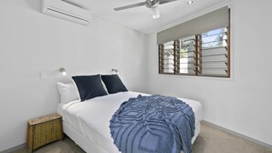Quarto com cama, ventilador de teto, ar condicionado, mesa de cabeceira e janela com persianas.