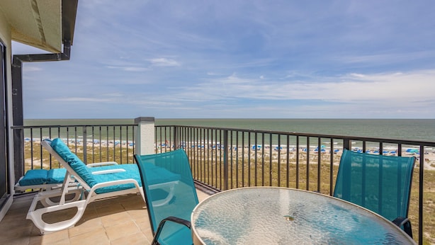 A balcony with two lounge chairs and a glass table, overlooking a beach with umbrellas and the ocean.