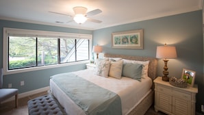 A bedroom with a large bed, two bedside lamps, a ceiling fan, a window with a view, and a small bedside table with a lamp and picture frame. - Hilton Head Villa: 2 King Suites, Loft, Updated Interiors & Beach Perks (Hilton Head Island)