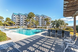 Área da piscina com espreguiçadeiras, uma pérgola de madeira e um edifício com varandas.