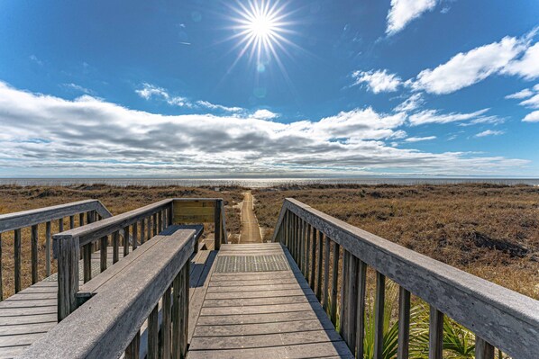 A wooden boardwalk leading to a beach with a clear sky and sun.
