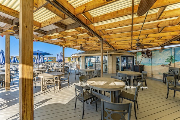 Un'area salotto esterna con tavoli e sedie in legno, una ventola a soffitto e un murale raffigurante una spiaggia.