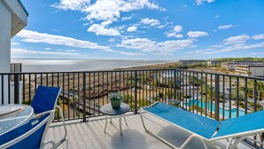 A balcony with a view of the beach, a pool, and a dining area with chairs and a table.