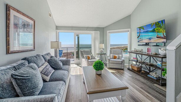 A spacious living room with a gray sofa, a wooden coffee table, and a TV on a wooden stand. There is a balcony with a view of the ocean.