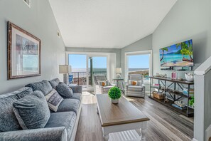 A spacious living room with a gray sofa, a wooden coffee table, and a TV on a wooden stand. There is a balcony with a view of the ocean.