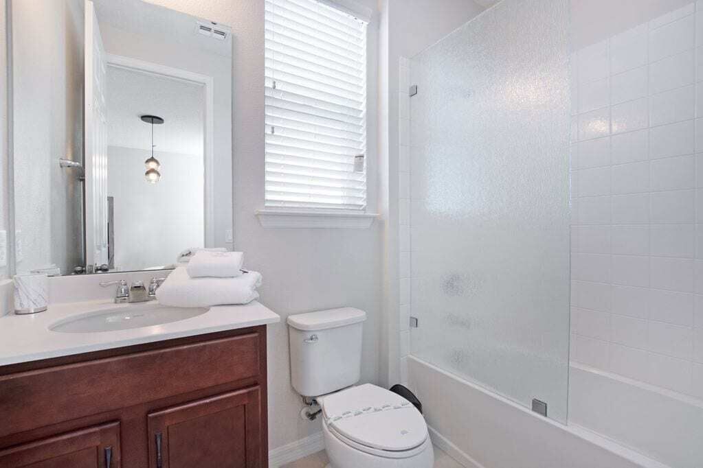 A bathroom with a white sink, mirror, and toilet, a shower with frosted glass doors, and a window with blinds.