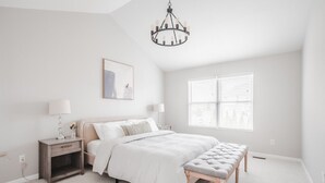 A bedroom with a bed, bedside tables, a bench, a chandelier, a window with blinds, and a wall-mounted picture.