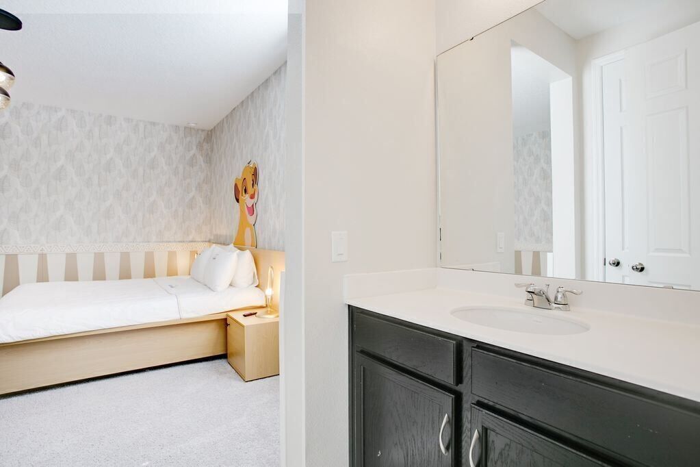 Un dormitorio con cama, mesita de noche y un mueble con lámpara. El baño tiene lavamanos, espejo y armarios.