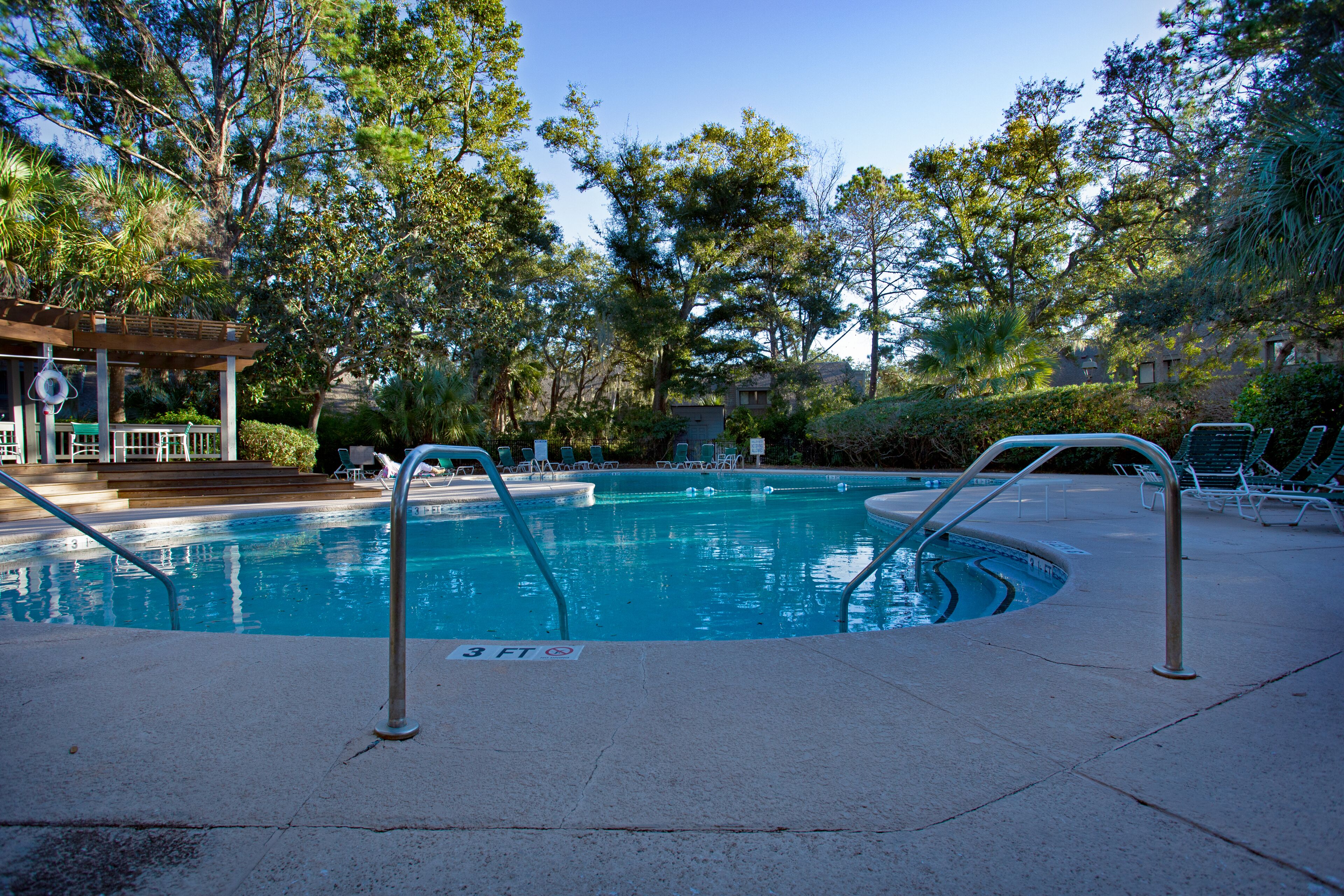 Une piscine dont la profondeur maximale est de 3 pieds, entourée d’arbres et de chaises longues.