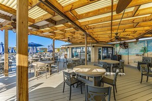 An outdoor patio with wooden tables and chairs, umbrellas, and a mural of a beach scene.