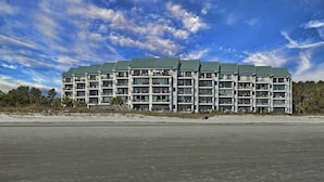 Un hotel de varios pisos con techo plano y balcones, situado cerca de una playa bajo un cielo despejado.