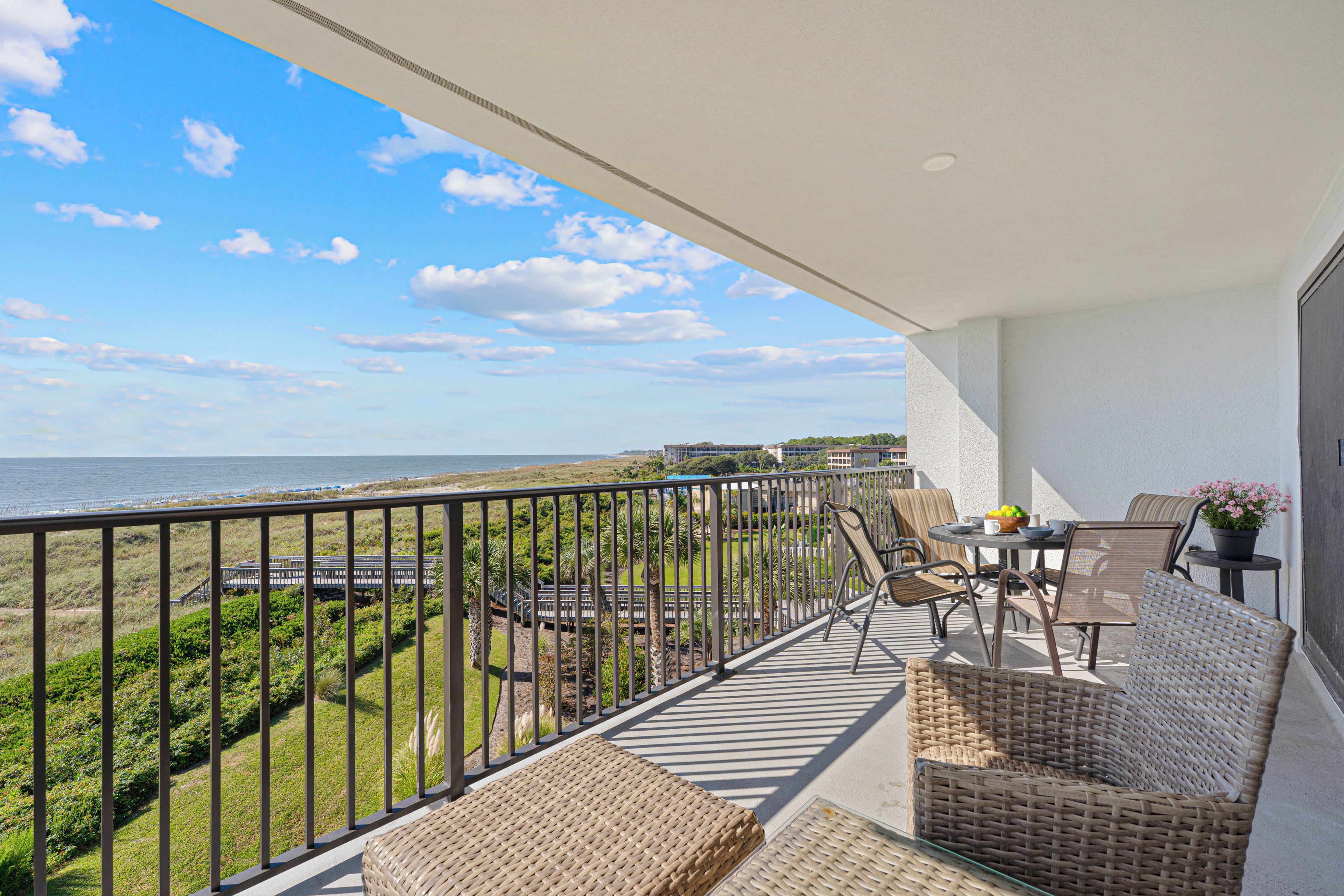 A balcony with wicker furniture, a table with a bowl of fruit, and a view of the beach and ocean.