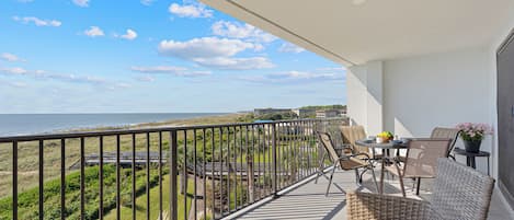 A balcony with wicker furniture, a table with a bowl of fruit, and a view of the beach and ocean.