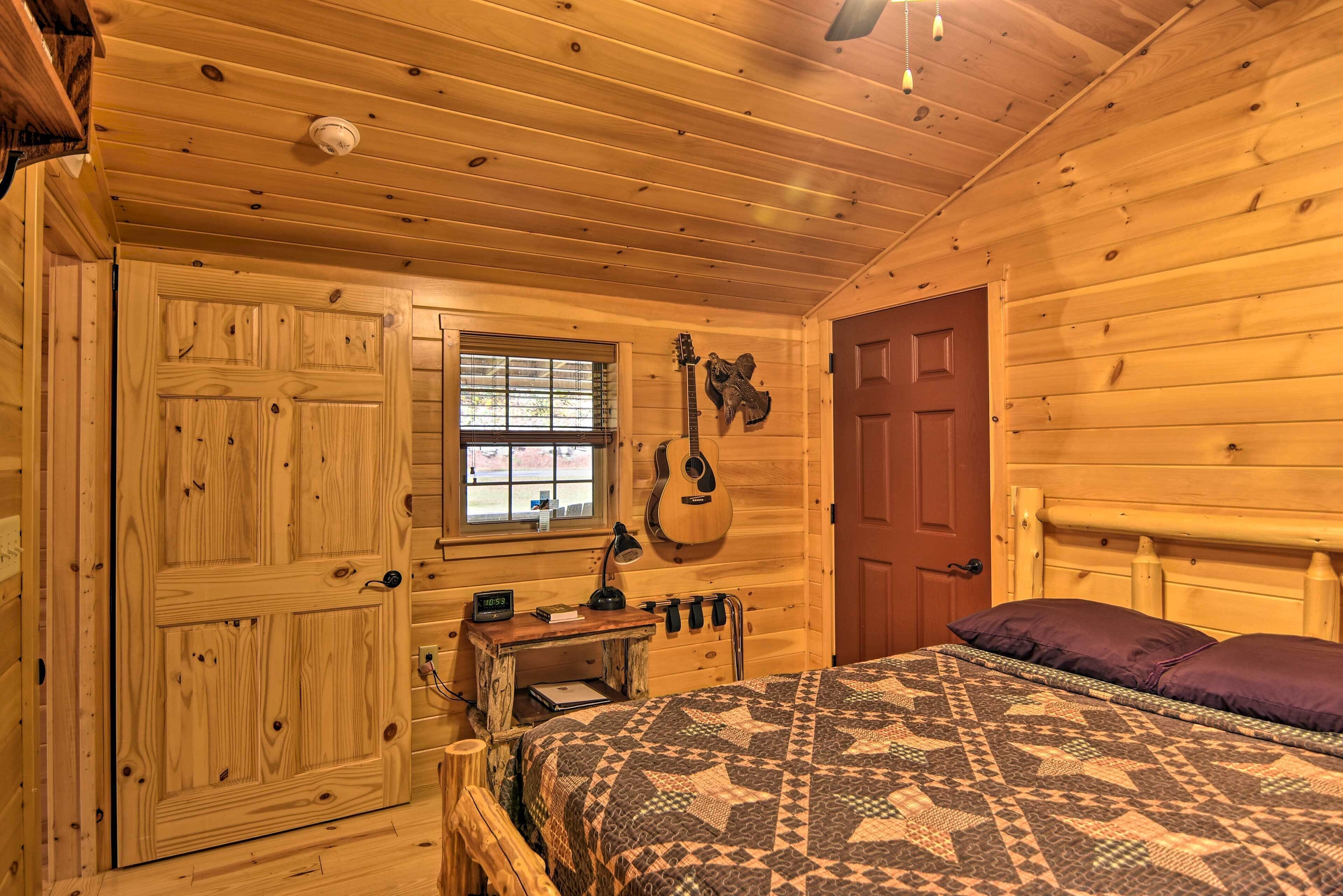 A wooden cabin bedroom with a bed, a desk, a guitar, and a window.