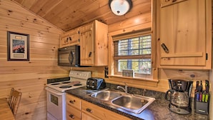 A wooden cabin kitchen with a stove, sink, and a window with a view of trees.