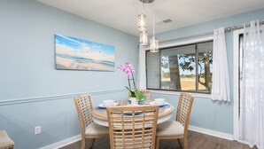 A dining area with a round table, four chairs, a painting of a beach scene, a potted plant, and a window with curtains.