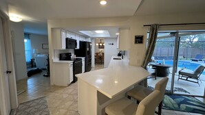Eine moderne Küche mit weißen Schränken, einem schwarzen Kühlschrank und einer großen Kochinsel. Es gibt einen Essbereich mit Tisch und Stühlen sowie einen Blick in einen Garten mit Pool und Liegestühlen.