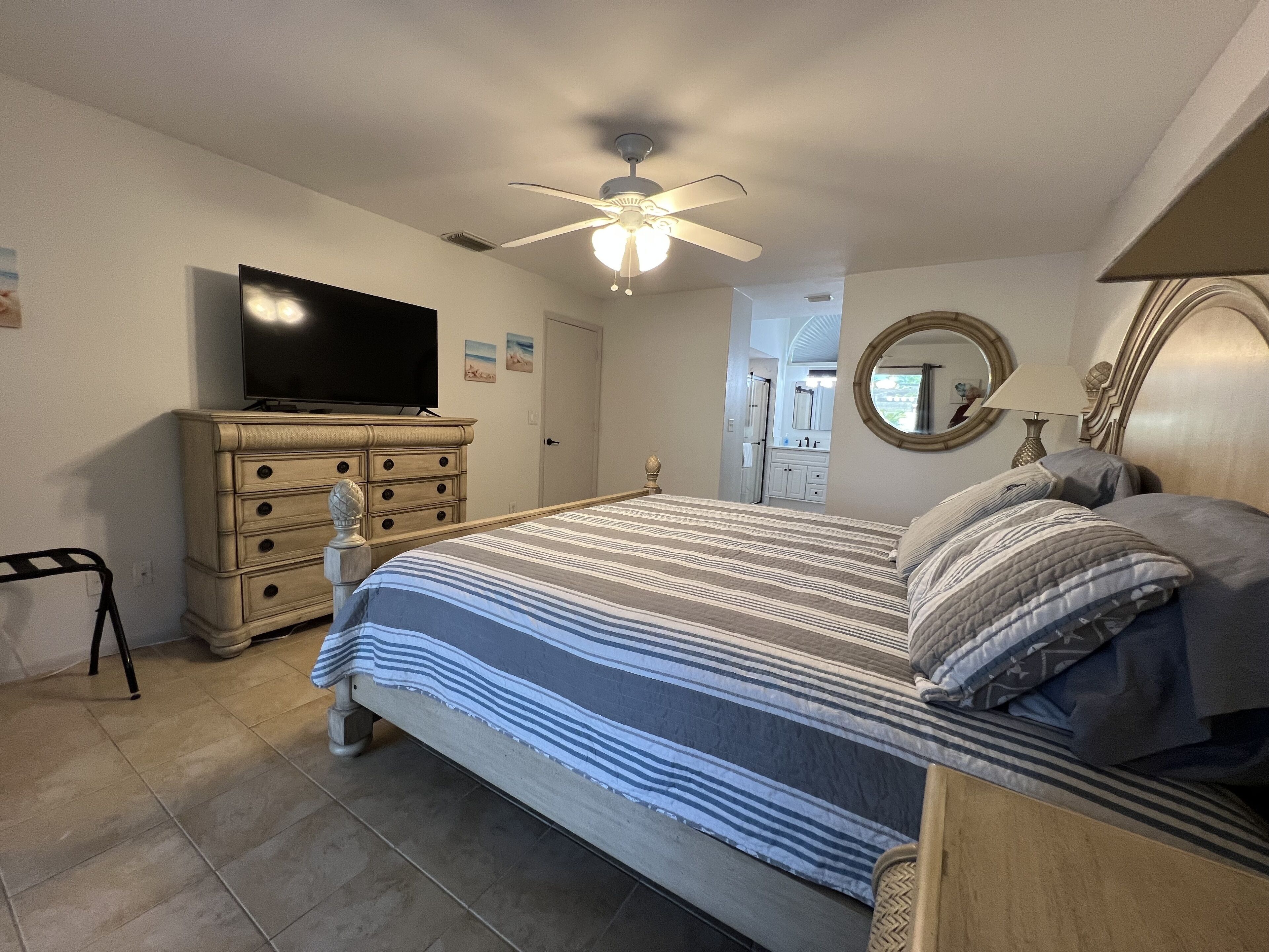 A bedroom with a bed, dresser, television, ceiling fan, and a bathroom visible through an open door.