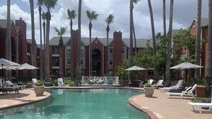 A pool area with lounge chairs and umbrellas, surrounded by tall palm trees and a brick building with arched windows. - Brand New near Houston Medical Center / Rice University / NRG Stadium (Houston)