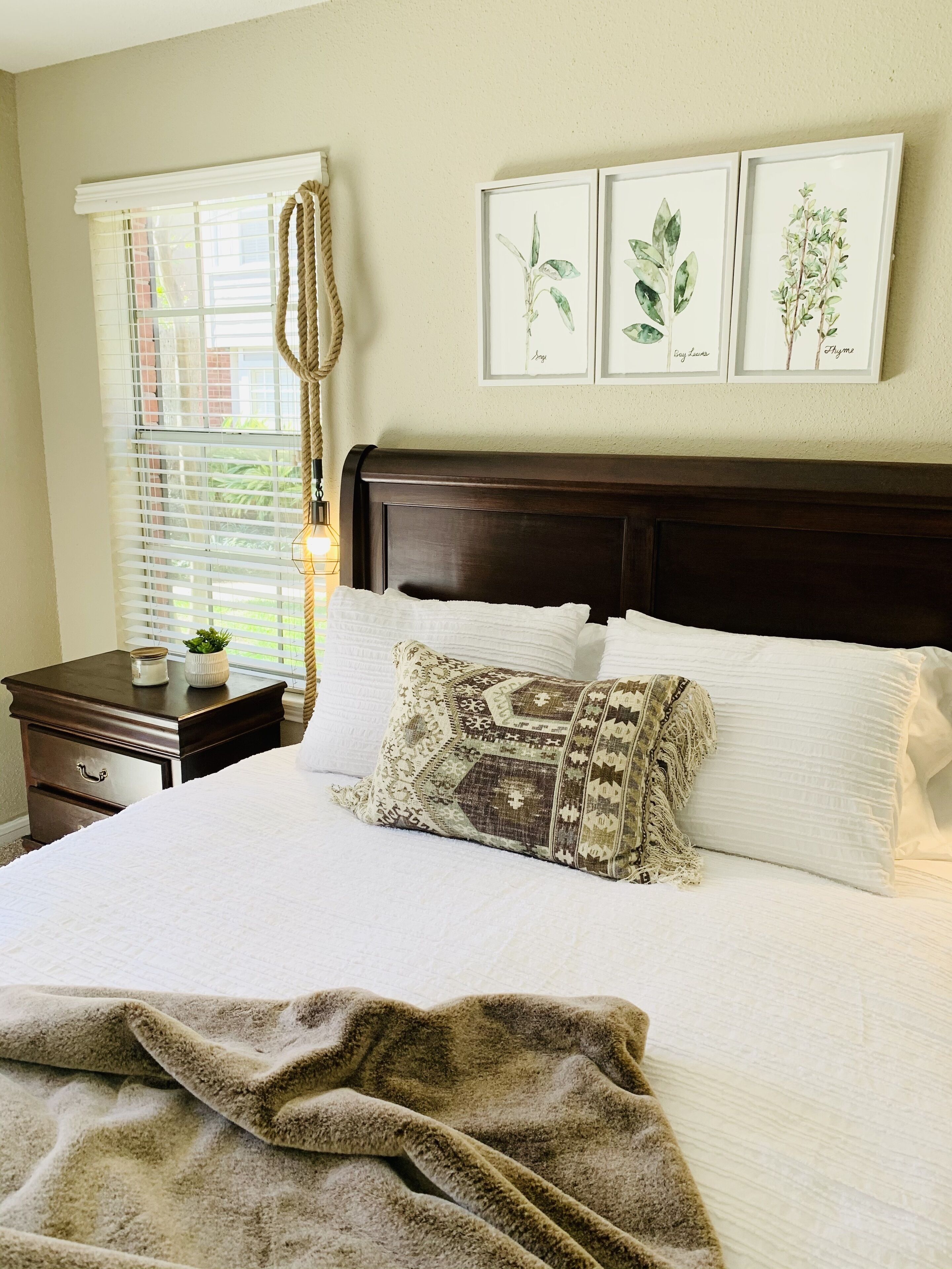 A neatly made bed with a patterned pillow and a blanket, a bedside table with a plant, and framed botanical prints on the wall.