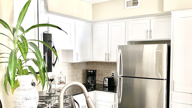 A modern kitchen with white cabinets, a stainless steel refrigerator, a coffee maker, and a large green plant. - Brand New near Houston Medical Center / Rice University / NRG Stadium (Houston)